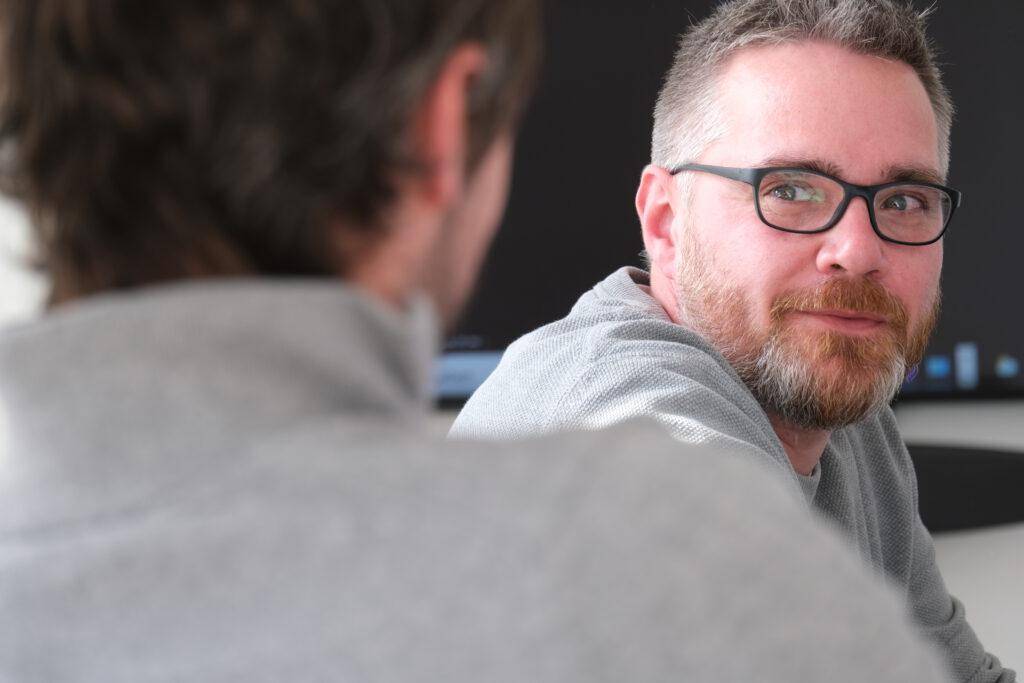 Workshop Rabot Energy: Mann mit Brille und Bart sitzt in einem Konferenzraum und schaut zu einem anderen Mann, der im Vordergrund verschwommen zu sehen ist.