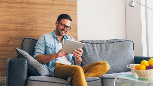 Ein Mann sitzt auf einem Sofa und nutzt ein Tablet, während er lächelt.