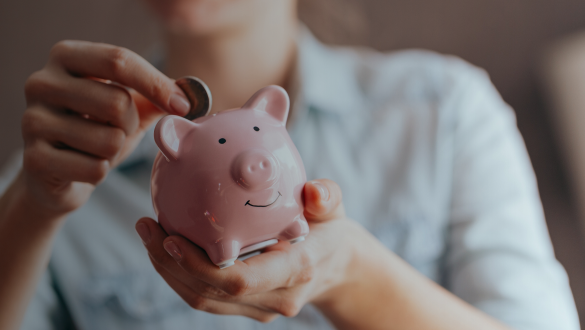 Eine Person hält ein rosa Sparschwein in den Händen und steckt eine Münze hinein. Das Sparschwein hat ein freundliches, lächelndes Gesicht. Im Hintergrund ist die Person unscharf zu sehen, die eine helle Bluse trägt. Eine Person hält ein rosa Sparschwein in den Händen und steckt eine Münze hinein. Das Sparschwein hat ein freundliches, lächelndes Gesicht. Im Hintergrund ist die Person unscharf zu sehen, die eine helle Bluse trägt.