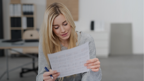 Eine junge Frau sitzt an einem Schreibtisch und liest aufmerksam ein Blatt Papier. Sie hält einen Stift in der Hand, bereit, Notizen zu machen. Im Hintergrund ist ein modernes Büro mit Regalen und einem Computerarbeitsplatz zu sehen.