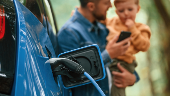 Mann lädt sein E-Auto mit günstigem Öko Strom und hält dabei seinen Sohn auf dem Arm.